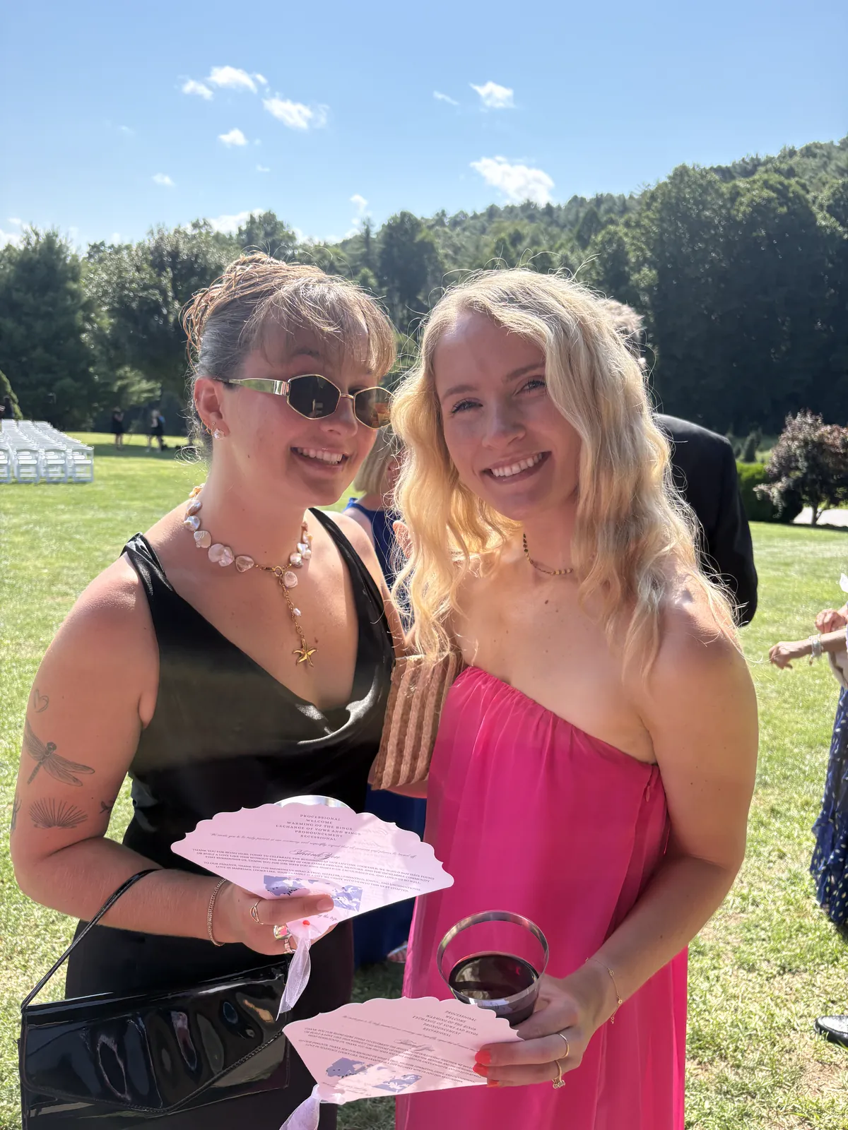 Kaitlyn in a pink dress with a friend, both holding paper fans on a sunny lawn