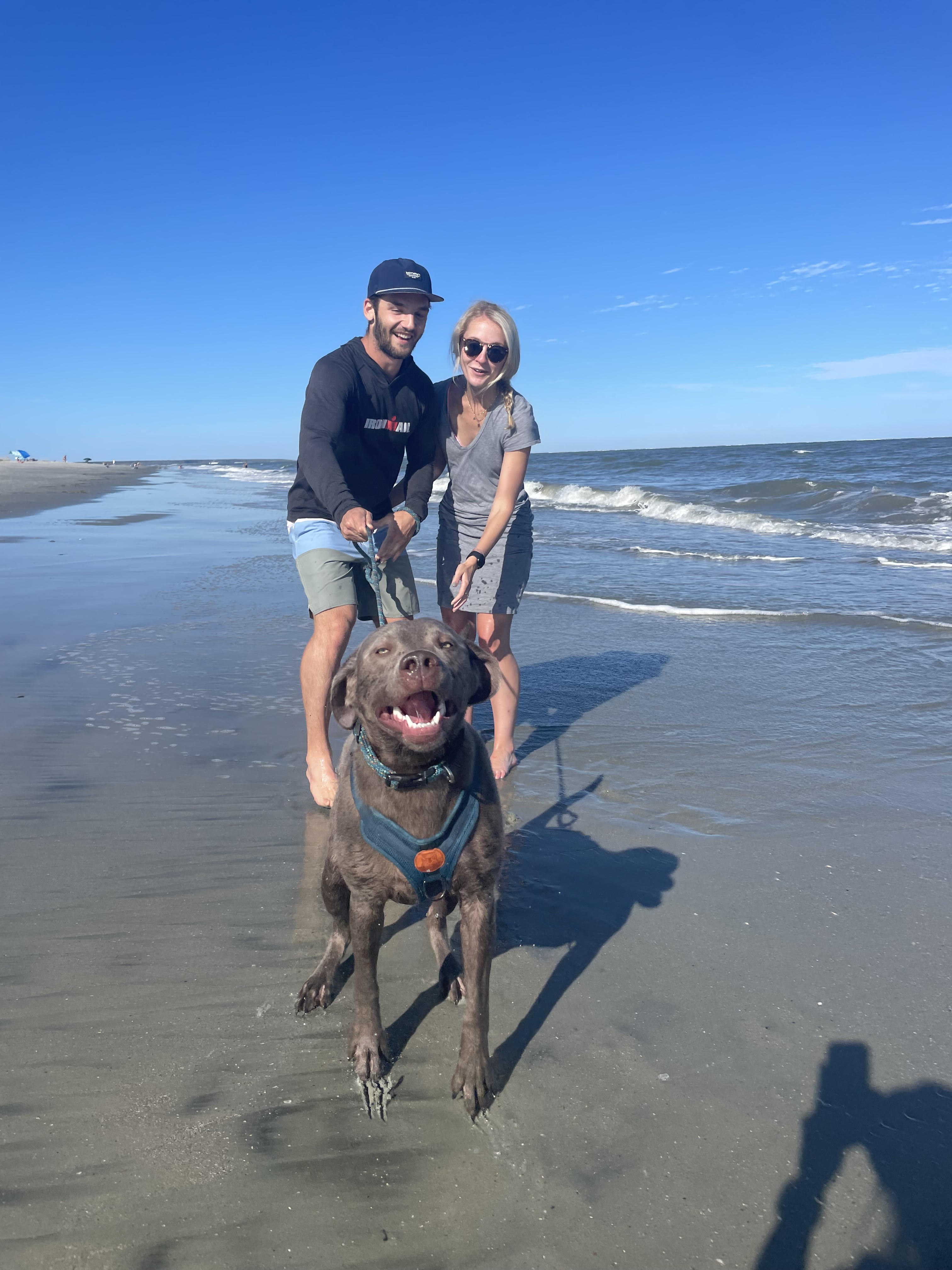 Maverick grinning at the camera, with Kaitlyn and Jack holding his leash on the beach behind him.