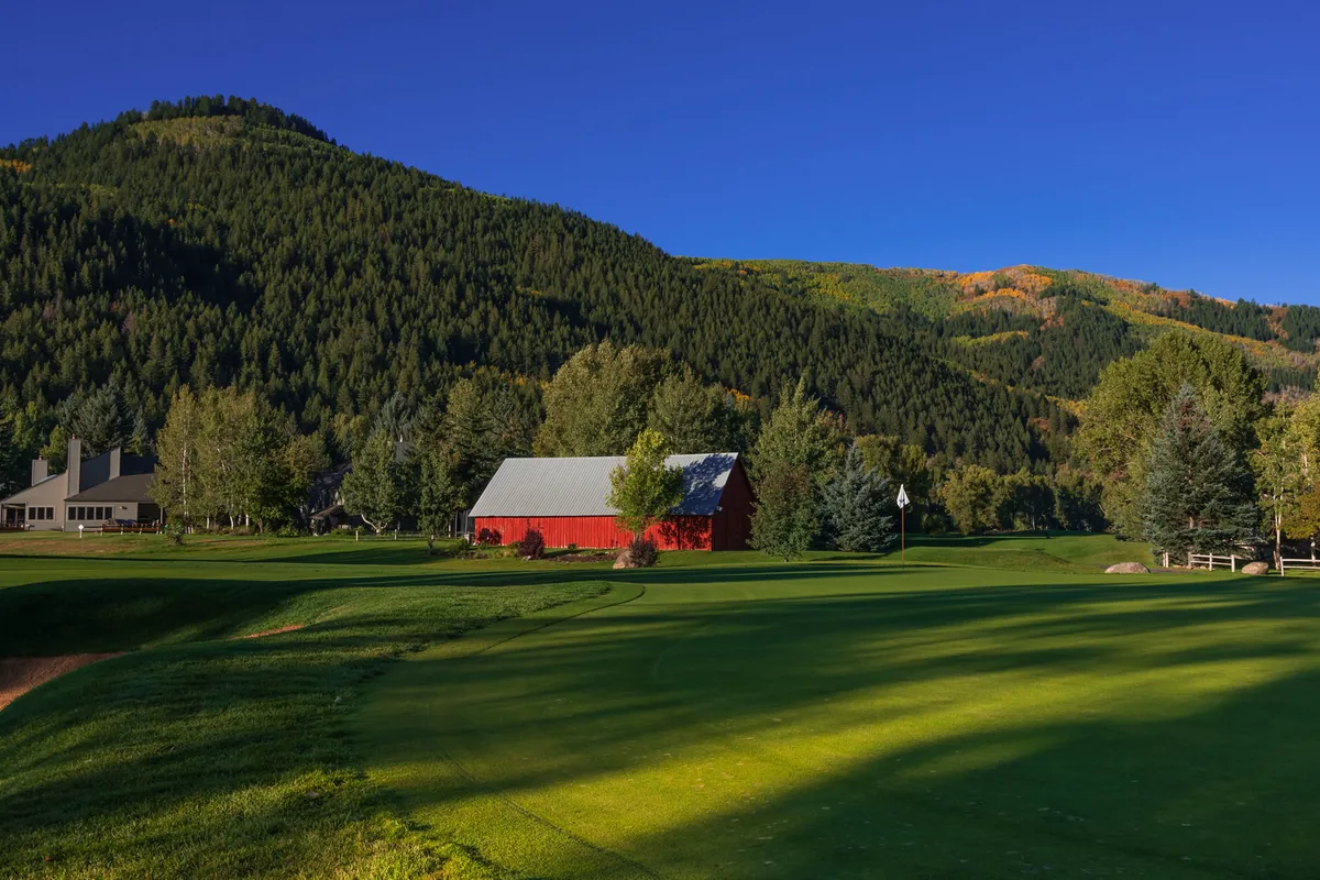 A sunlit fairway at Eagle-Vail Golf Club with a red barn below a forested mountain